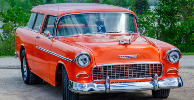 An orange 1955 Chevy Nomad parked in-front of a green forest area.