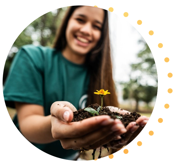 Person holding a small mound of soil with a blooming yellow flower and seedling outdoors, background blurred