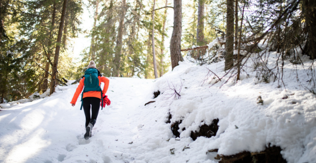A woman, wearing an orange coat with gloves attached to her blue backpack is taking strong strides up hill. There is snow covering the ground. The sun can be seen through the trees in the forest.
