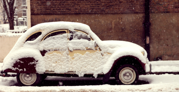 A collector car covered in snow is parked on the side of a road in front of a building with a red brick wall.