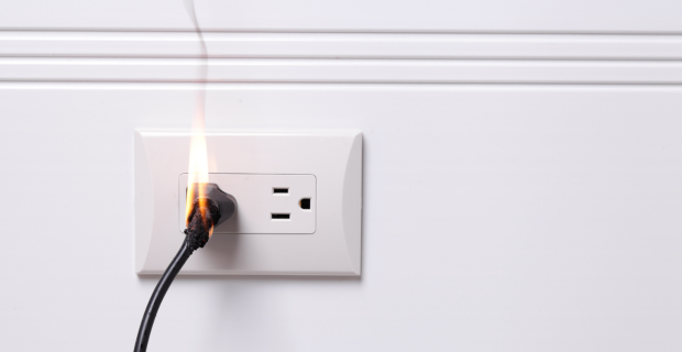 Electrical plug on a white wall has a small fire coming out of it. 