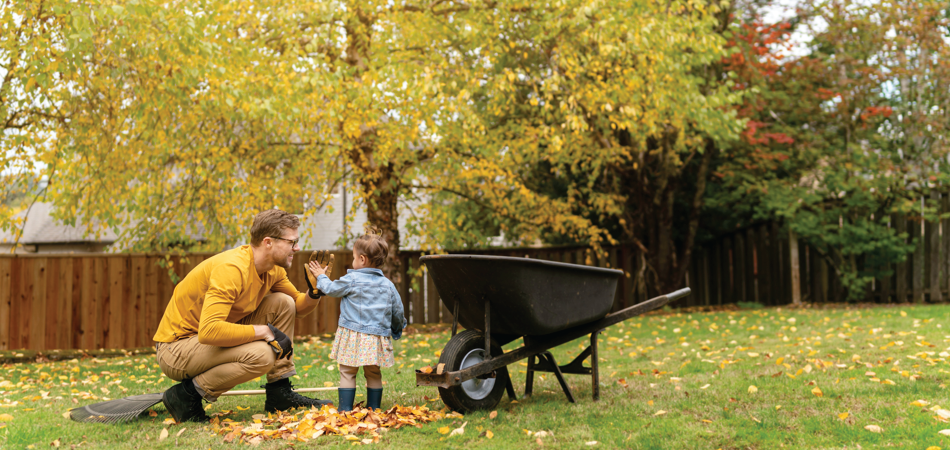 A person and a child in a yard with autumn leaves beside a wheelbarrow.