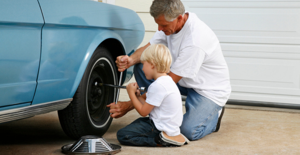 A man in a white shirt and denim jeans is showing a young child in a white shirt and denim jeans how to use a tire jack. A hubcap is on the ground beside the child. In front of them is a stunning baby blue classic car.