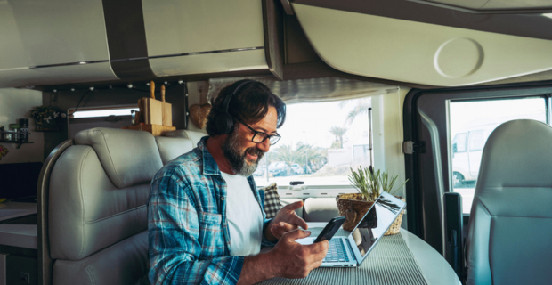 A man appears to be in a video call. He is smiling as he holds his phone, his hands are animated, appearing to have an exciting conversation. On his RV tabletop is an open laptop. The interior of the RV is cosy and modern with tan seating.