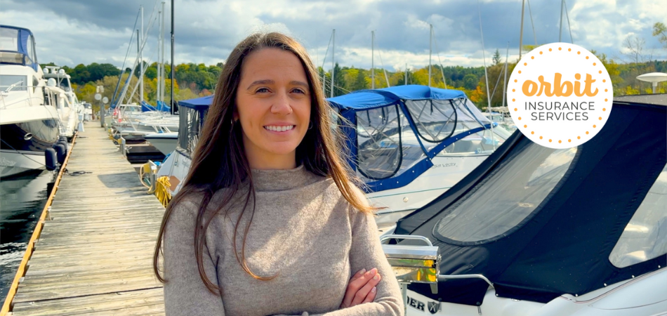 A person with long hair stands on a dock talking about marine insurance. They’re smiling and wearing a beige sweater while several boats are docked behind them.