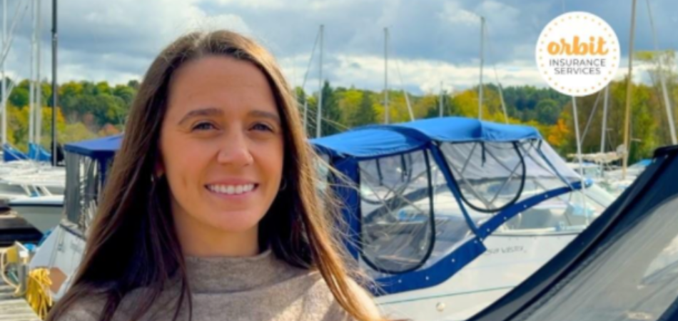 A person with long hair stands on a dock talking about marine insurance. They’re smiling and wearing a beige sweater while several boats are docked behind them.
