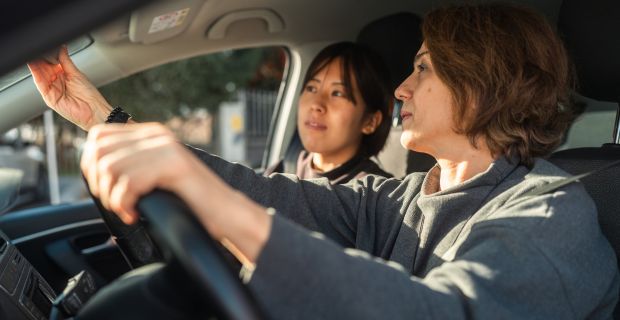 Two women sitting in a car, the driver is adjusting their rear view mirror.