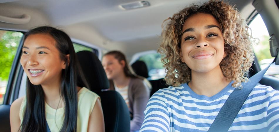 Young person driving a car with two passengers, all wearing seatbelts, travelling together on a sunny day.