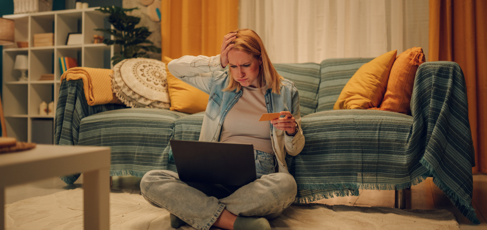 A person sits on the floor of their living room, holding a credit card and looking worriedly at their laptop, suggesting a possible encounter with an online shopping scam.