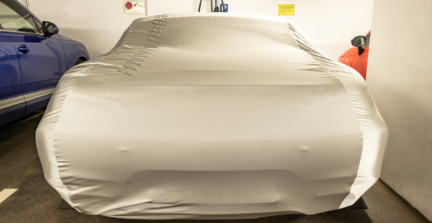 A collector car is parked in a garage between a blue and an orange car. Th collector car is covered with a quality car cover that is tan in colour