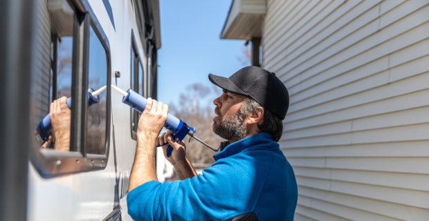 A man is using a caulking gun to fix his outside window frame on his trailer. His trailer is parked close to a house with siding.