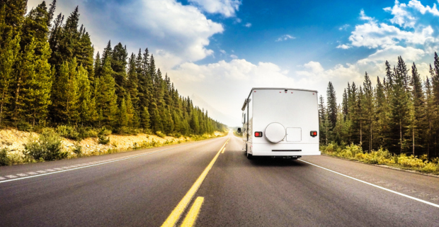 The back of an RV can be seen driving down an open highway. The sky is brilliant blue with some fluffy clouds floating. The highway is in the middle of a beautiful, forested area.