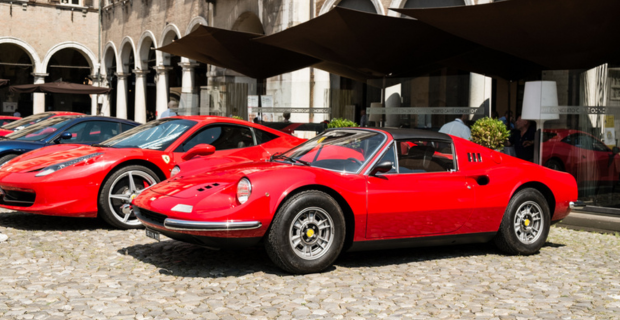 A row of gorgeous red Ferrari’s and a stunning blue Ferrari are parked in front of a mirrored building with arches and outdoor umbrellas. They are parked on a cobblestone road