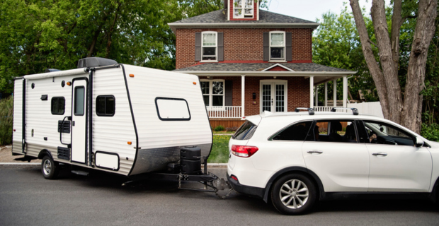 A white four door car is pulling a white trailer leaving home, on the way to an adventure! In the background a red brick house with a wraparound porch can be seen with a large tree in the front yard.