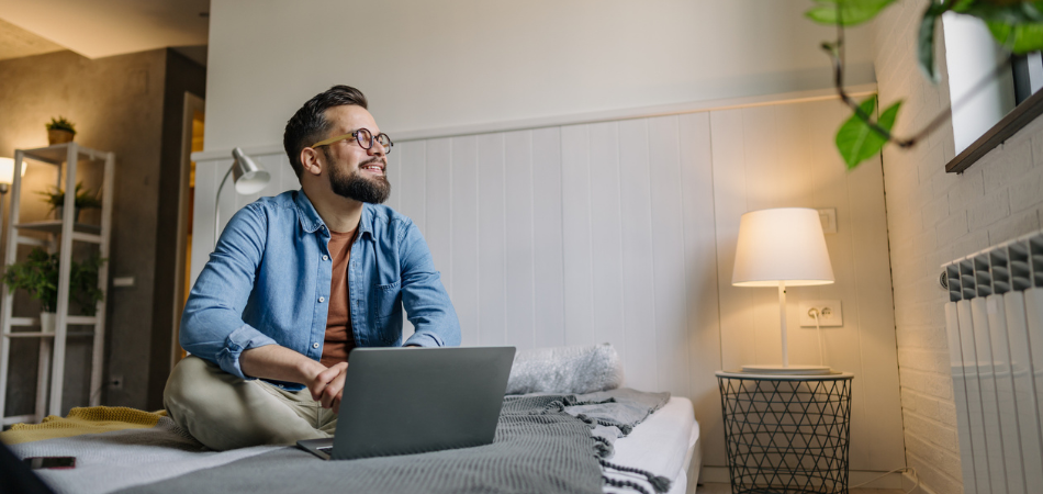 Young man in his apartment researching renters insurance on his laptop while sitting comfortably on his bed in a cozy, well-lit bedroom.