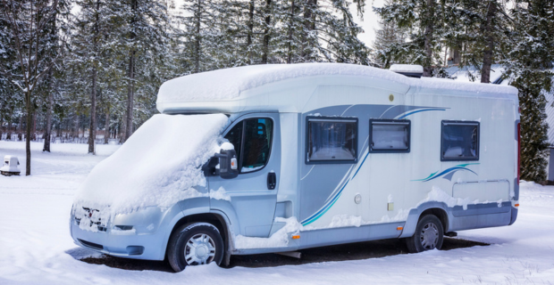 A stunning blue and white RV is seen parked outside covered in snow. There are trees and lots of snow on the ground.
