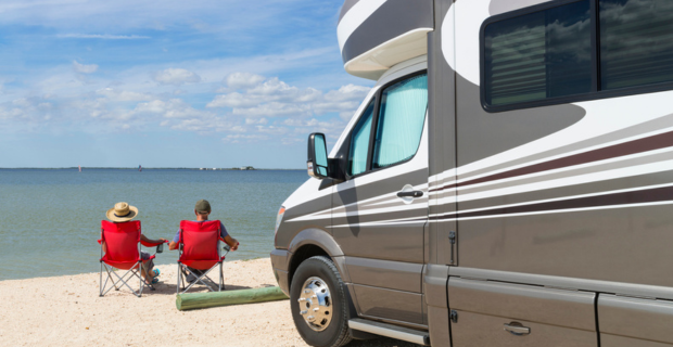 a modern RV is parked on a sandy beach. In the distance you can see the beautiful blue water. Sitting close to the water are two people soaking up the sun as they relax in red beach chairs