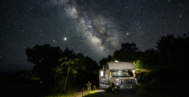 Under a spectacular night sky full of stars, a camper is set up in the dark, a picnic table is holding the makings for a late night meal.