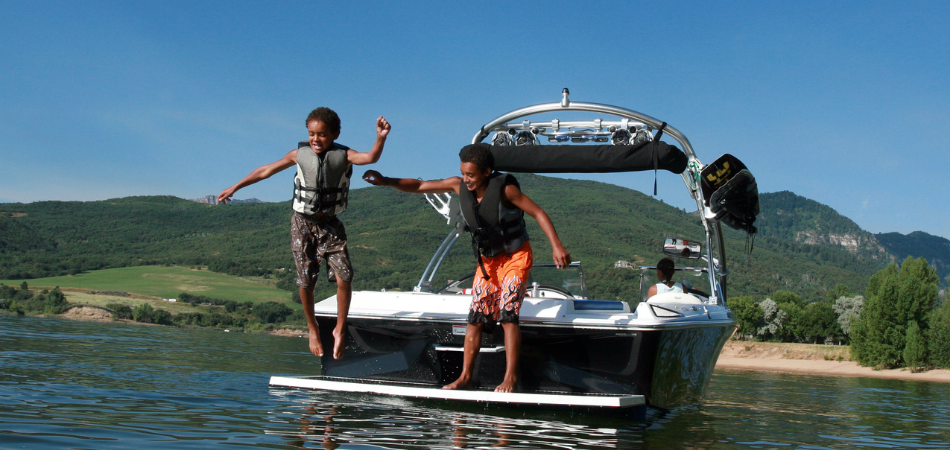 Two young boys seen jumping off a wake boat on a lake with life jackets on.  