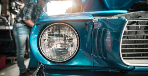 A close up image of a stunning blue collector car with a focus on the double headlight. A man in a plaid shirt, a hat, and denim can be seen working on the gorgeous car in the background