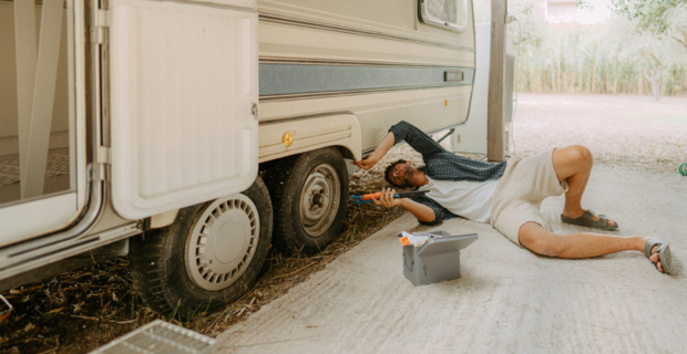 A man is wearing summer clothing, he is laying on the ground with a tool in his hand, he is fixing the tire of his trailer.