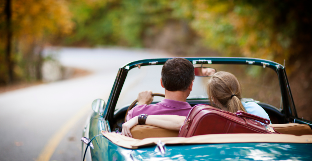 A couple is enjoying a drive on a beautiful warm day. The teal convertible collector car with tan interior is pristine. A burgundy suitcase can be seen in the backseat.