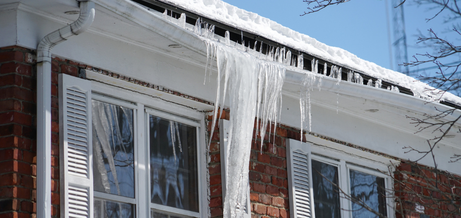 Icicles hanging from the edge of a snow-covered roof on a brick house with white shutters, under a bright blue winter sky.  