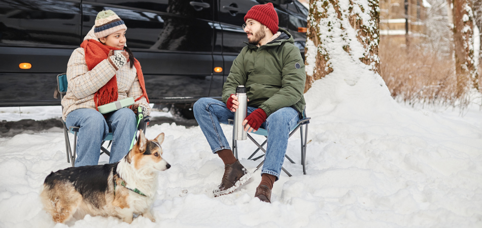 A couple sitting in chairs on a winter campsite with their dog in front of their motorhome.