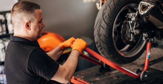 A man wearing a black shirt with a collar and light orange work gloves is hoisting a dark orange jack with the motorcycle on the lift. Only the tire can be seen with some shiny metal. The motorcycle is lifted, at eye level with the man working on the motorcycle.