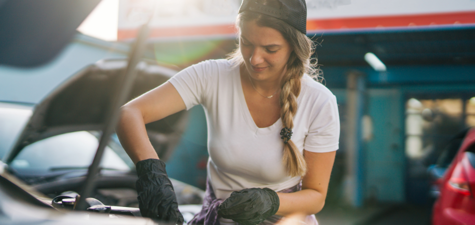 A young woman looking under the hood of a vehicle performing some classic car spring maintenance to get it ready to drive.