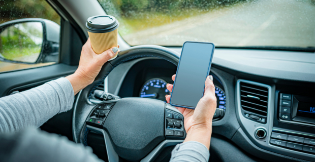A close-cropped photo of someone driving with their hand on the wheel, holding a coffee, with their phone in the other hand.