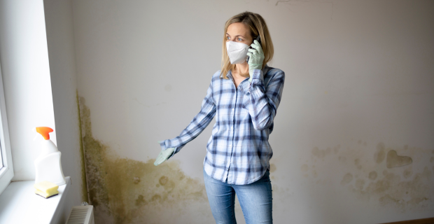 A Woman standing in a room with mould damage behind them wearing a mask while on the phone with her insurance company.