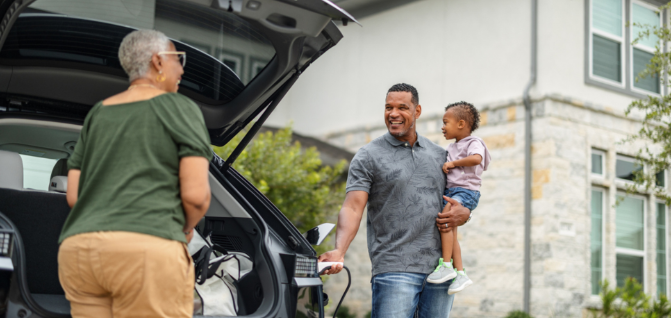 A person with short gray hair is unpacking their EV while another person holds a child and charges the vehicle. The car is parked in a residential area, and they’re talking about usage-based insurance.  
