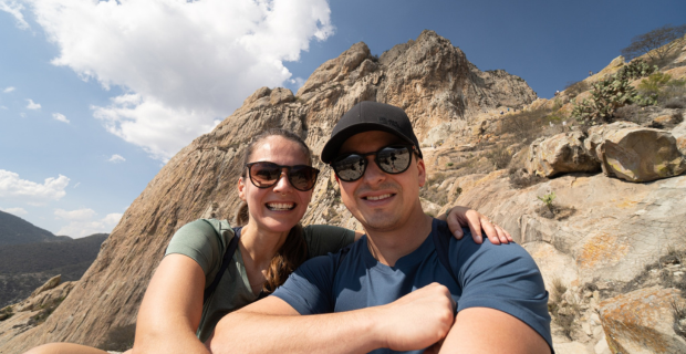Two people taking a photo. There are mountains in the background. It's a sunny day with some clouds in the blue sky.