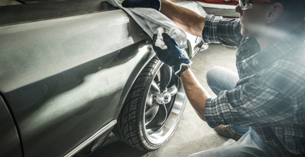 A man wearing denim and a plaid shirt is wearing a hat and safety glasses. He is wearing blue gloves and using a white towel to shine his prized collector car. Parked in front of his silver car that he is working on is a stunning red collector car