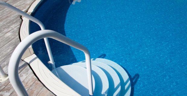 Looking down at a set of steps with handrails leading into an above ground pool surrounded by a wooden deck on a sunny day.
