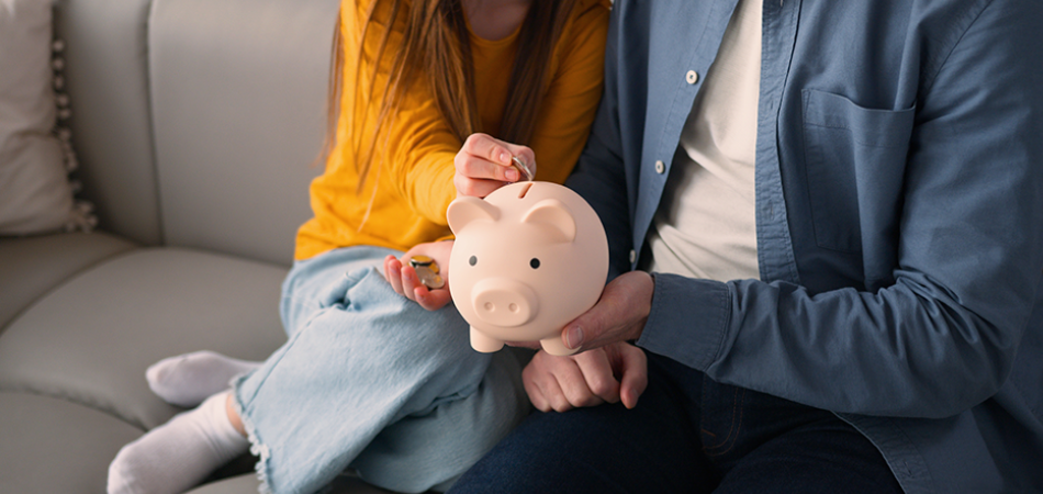 A young girl, with her father by her side, drops a coin into a pig‑shaped money bank.