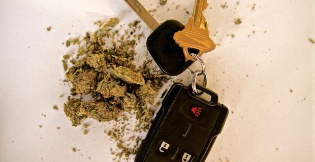 A set of car keys lie beside a small pile of dried cannabis on a white backdrop.