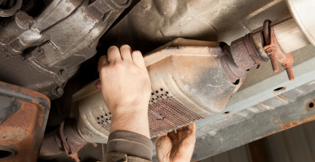 Two white hands reaching up to remove the catalytic converter from the underside of a well used, partially rusted vehicle. 