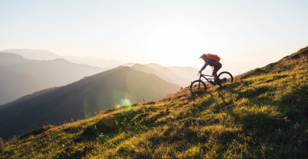 : A cyclist wearing a pack and riding down a grassy mountain. Beyond, a series of rolling mountains fade into the distance.