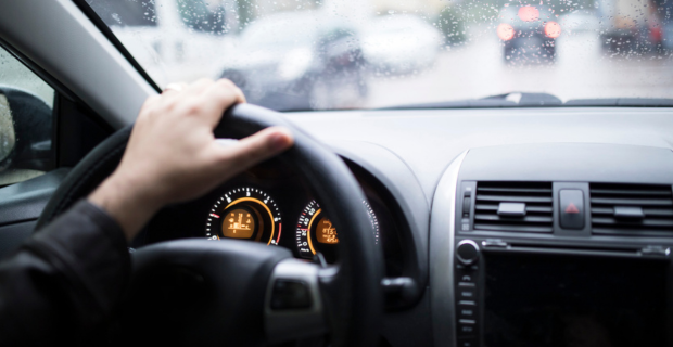 An out-of-focus hand at 12 o’clock on a steering wheel, through the wheel you can see the odometer and speedometer readings 