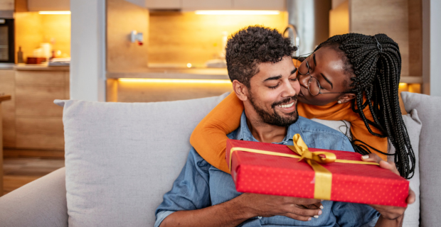 A Black man in a denim shirt relaxing on his couch. He is smiling as his partner, a Black woman with braids, wearing glasses, leans over his shoulder and passes him a gift wrapped in red paper and tied with gold ribbon. 