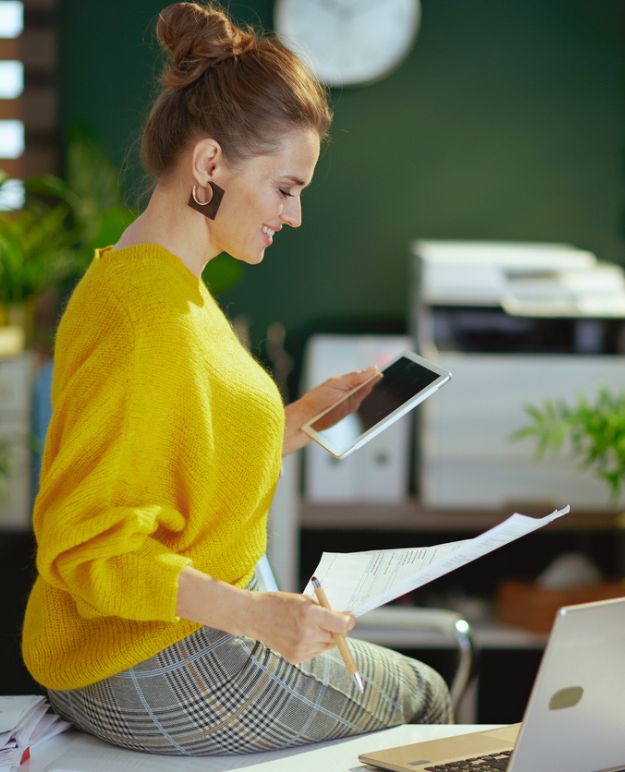 A woman in a yellow sweater sits on her desk reviewing accounts for her business.