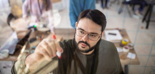 A heavyset man with glasses, a beard, and moustache is writing on a dry-erase board.