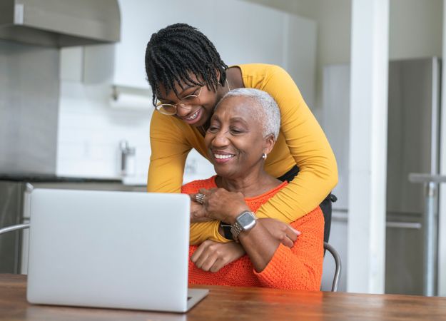 A Black teenager hugs her grandmother, who is working on a laptop at the kitchen table, from behind.