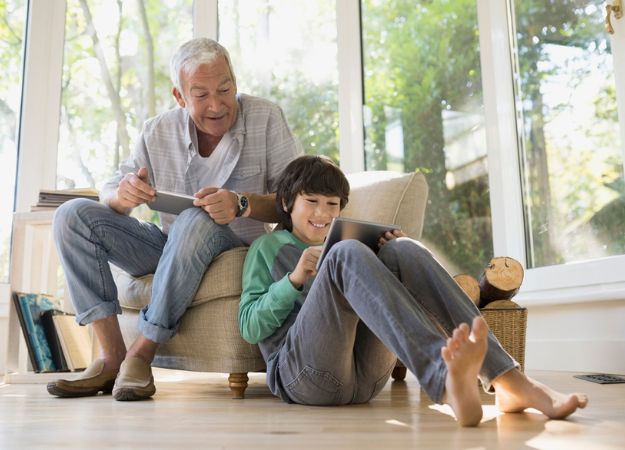 An Asian Canadian man sitting in an armchair watchin his grandson playing on a tablet.