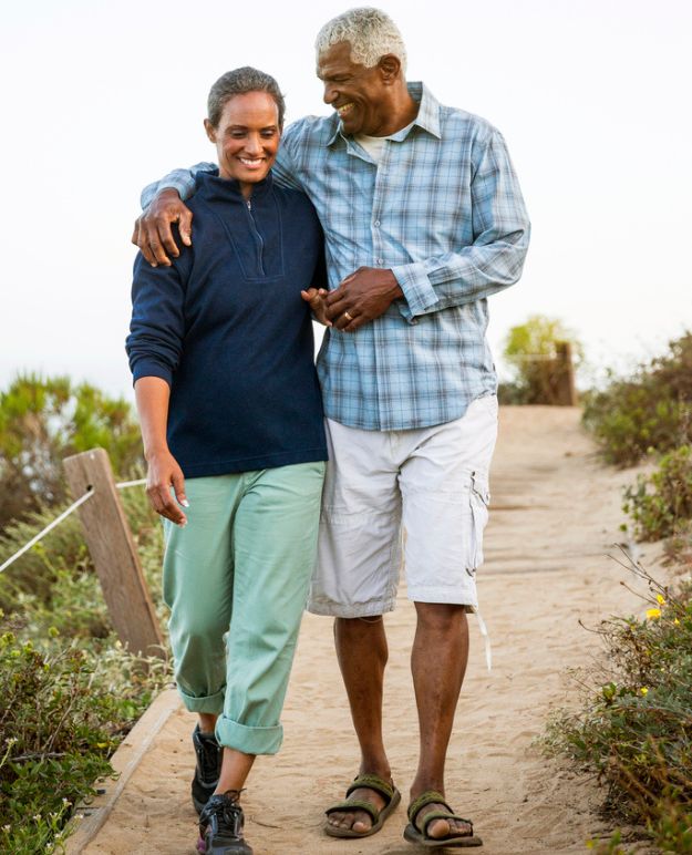 A couple in their 60s walking arm in arm along a boardwalk on a summer day.