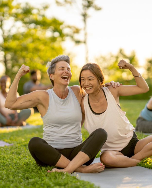 Two woman sitting cross-legged in a park doing yoga on a summer evening in Alberta. They are laughing, their arms around one another's shoulder, while they show off their biceps by flexing.
