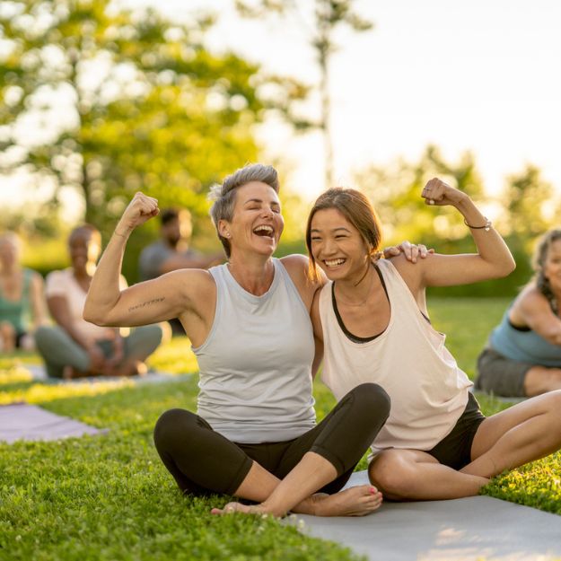 Two woman sitting cross-legged in a park doing yoga on a summer evening in Alberta. They are laughing, their arms around one another's shoulder, while they show off their biceps by flexing.