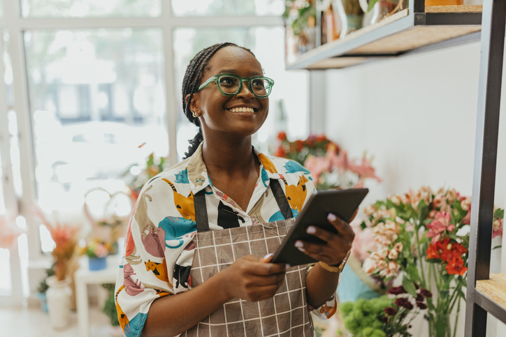 A smiling young woman wearing glasses holding a tablet in her flower shop